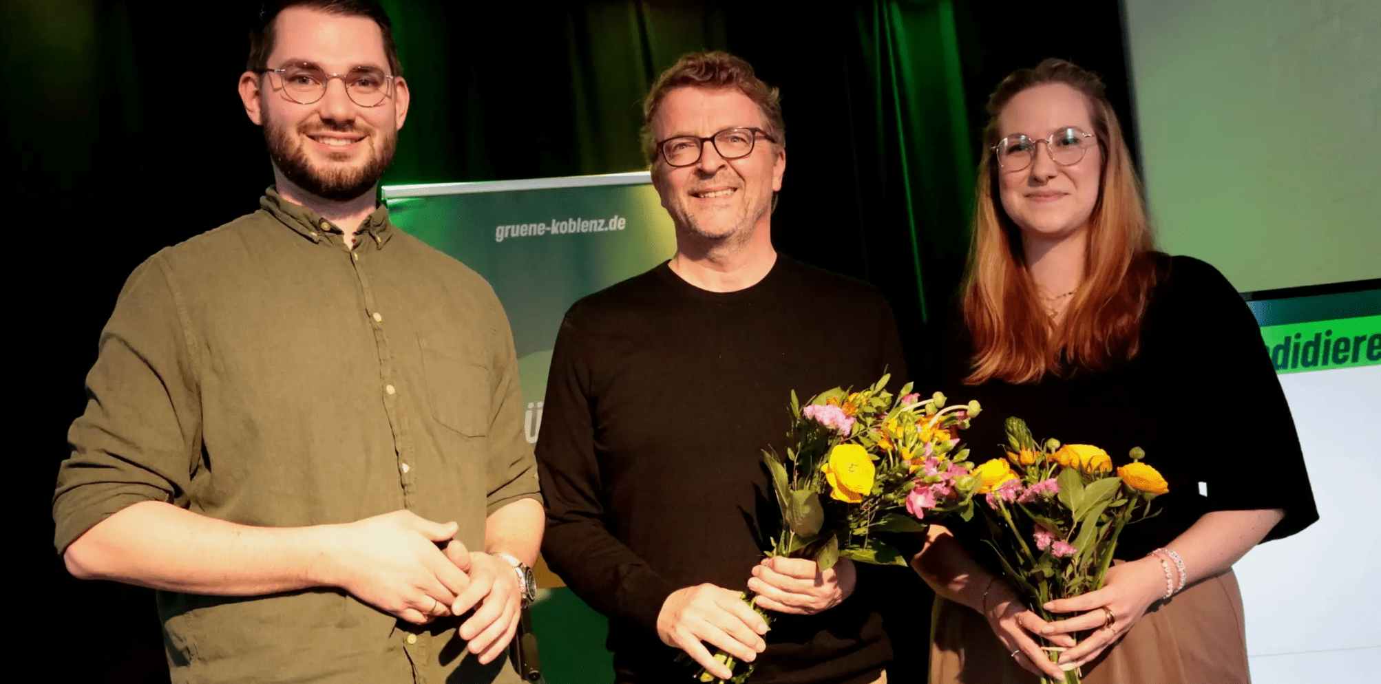 Christopher Bündgen, Carl-Bernhard von Heusinger und Janina Luipers stehen vor einem grünen Hintergrund und einem Plakat auf dem gruene-koblenz.de steht.