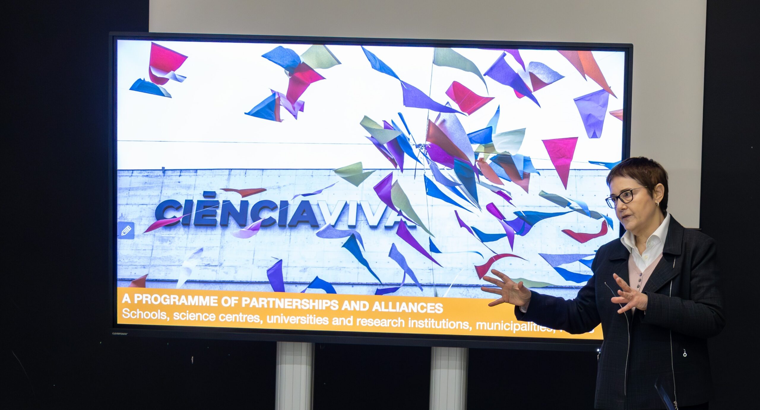 Dr. Ana Noronha vor einem Bildschirm der ein Bild des Centro Ciênca Viva Gebäudes zeigt. Unterschrift des Bilds lautet: A Programme of Partnerships and Alliances. Schools, science centres, universities and research institutions, munincipalities