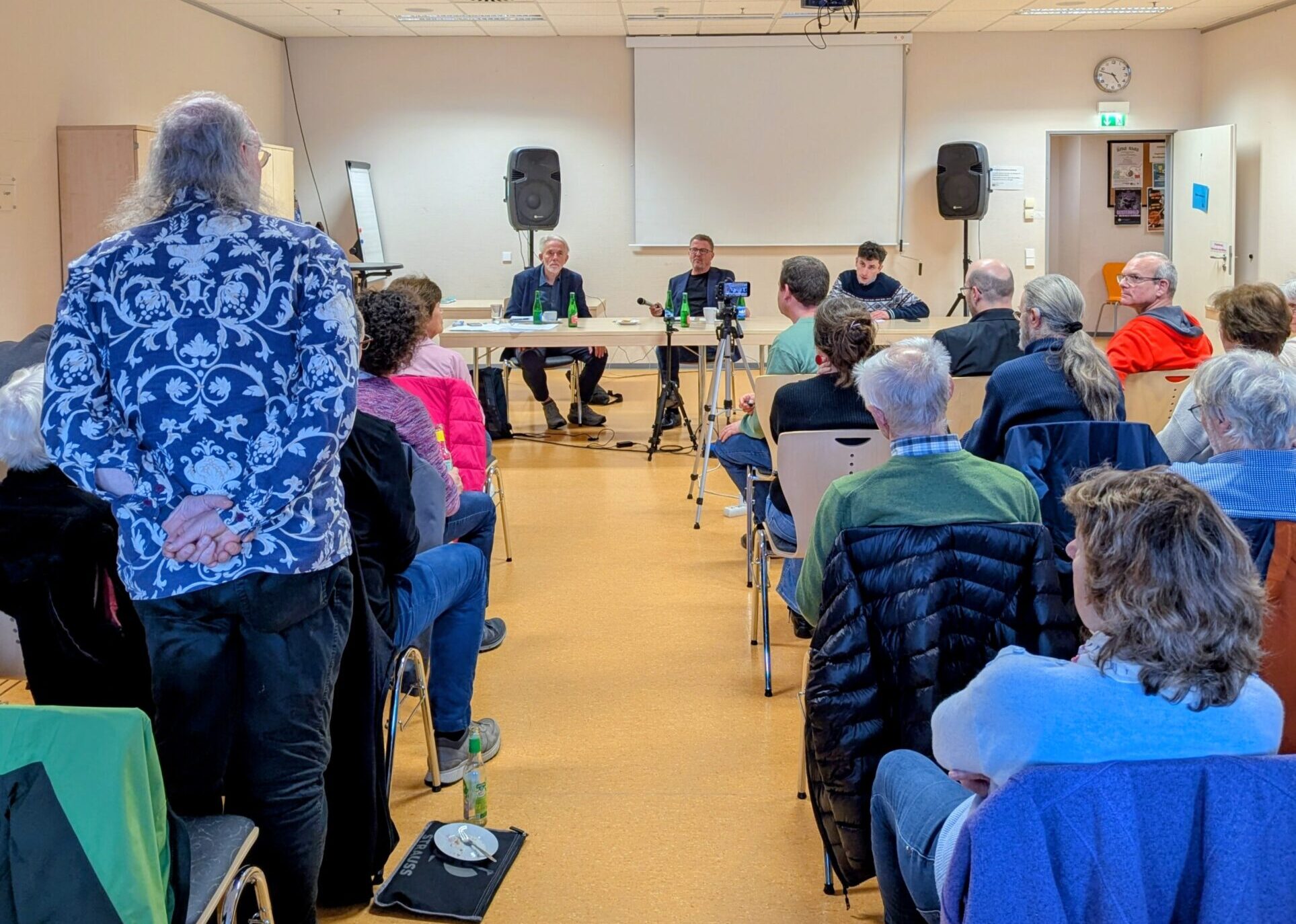 Foto vom Demokratieforum: Menschen sitzen auf Stühlen den Sprechern zugewandt, Prof. Dr. Rudolf Stichweh, Carl-Bernhard von Heusinger, Simon Schmitz sitzen nebeneinander an einem Tisch, dem Publikum zugewandt. Eine Person aus dem Publikum steht und die Sprecher gucken sie an.